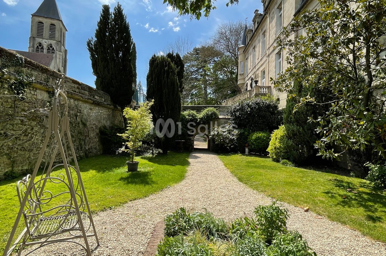 Jardin avec allée gravillonnée, végétation variée, bancs, et clocher d'église visible en arrière-plan.