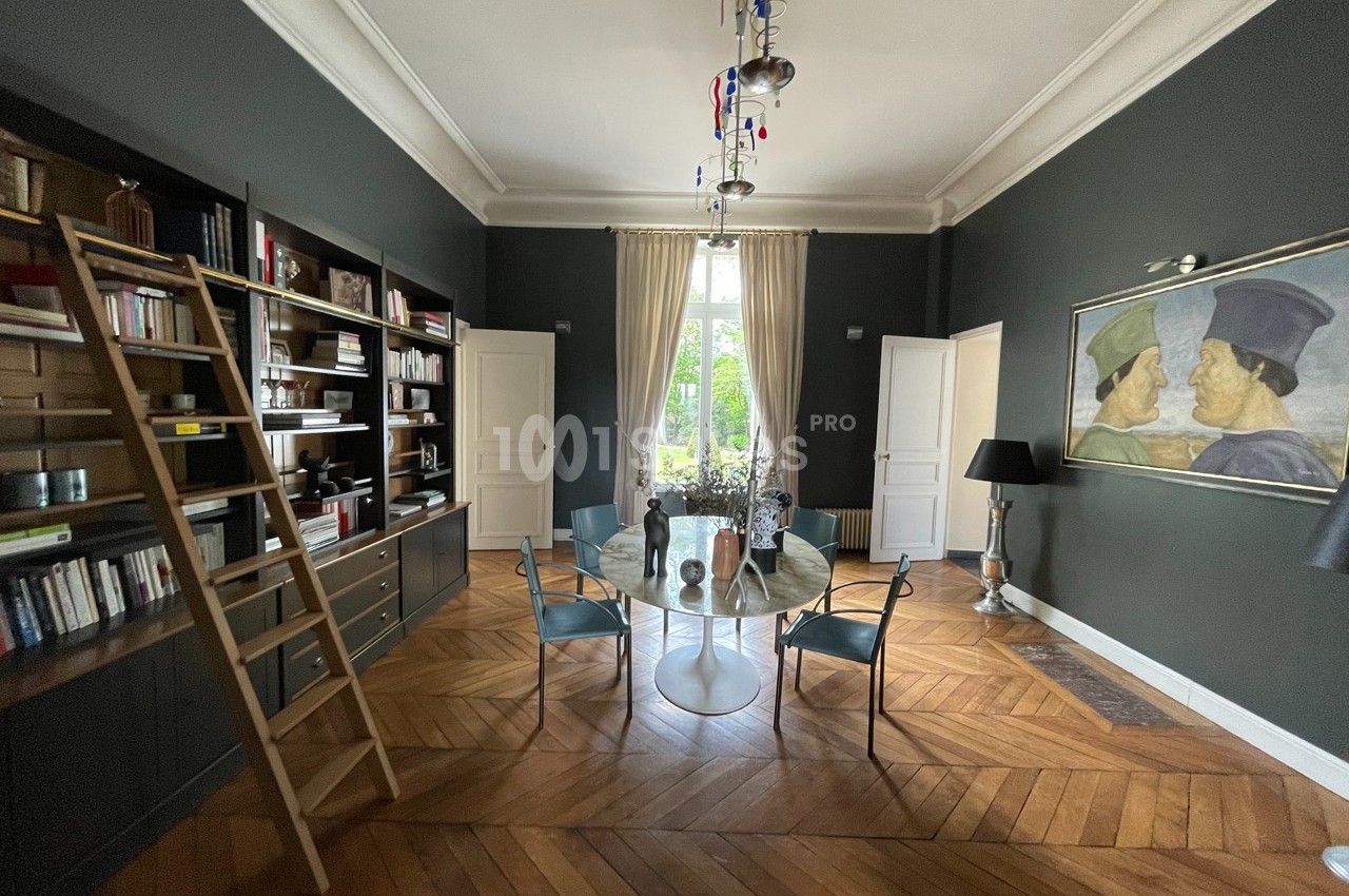 Salle à manger avec parquet en bois, table ronde en verre, étagères avec échelle, et grande fenêtre encadrée de rideaux.