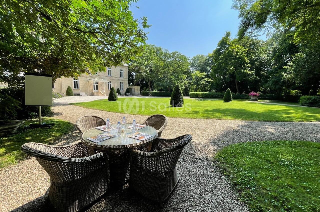 Table en rotin avec quatre chaises sur une allée gravillonnée, face à un jardin verdoyant et une grande maison en pierre.