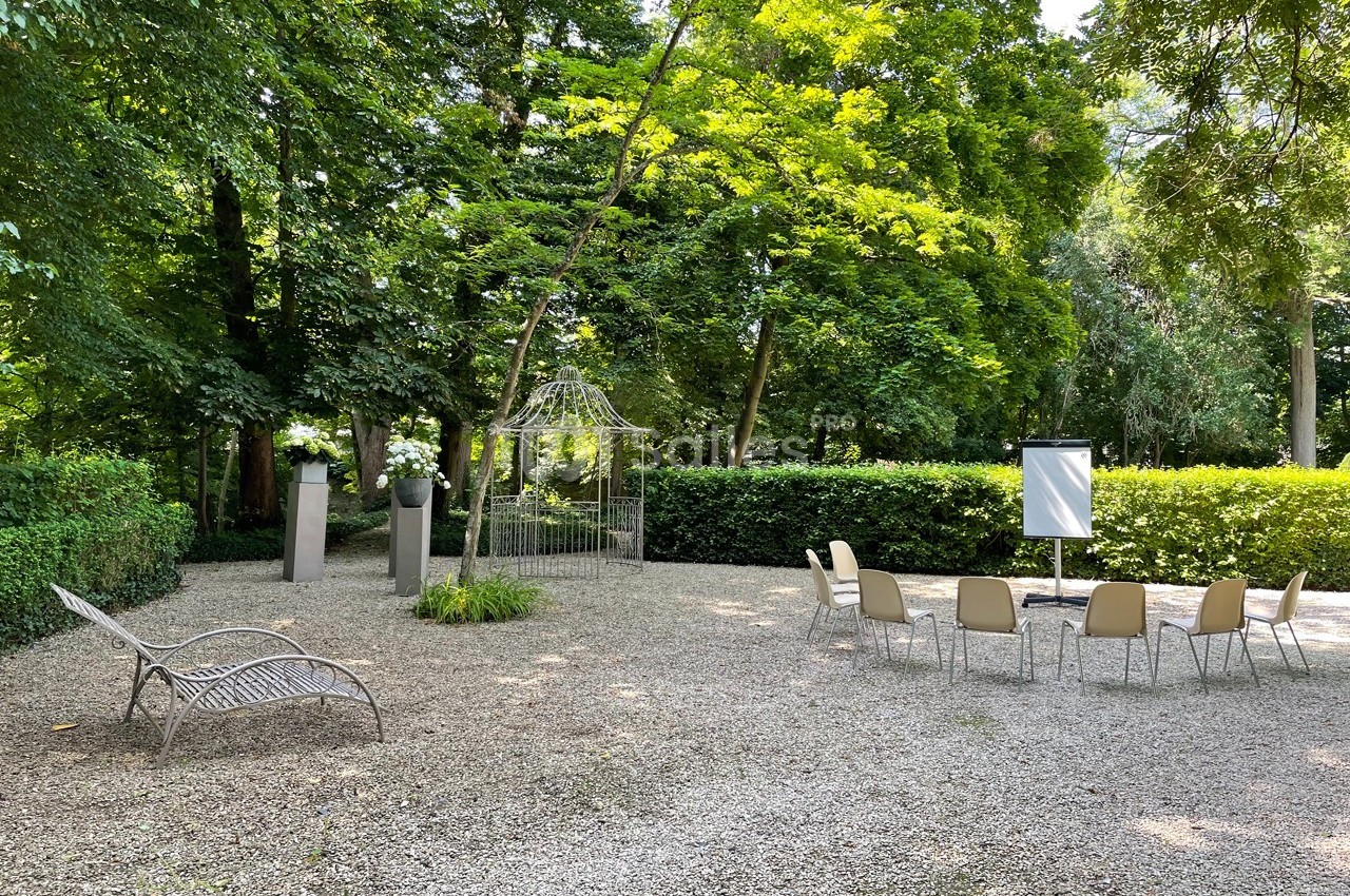Espace extérieur avec des chaises disposées en cercle, un chevalet blanc et une tonnelle entourée de verdure.