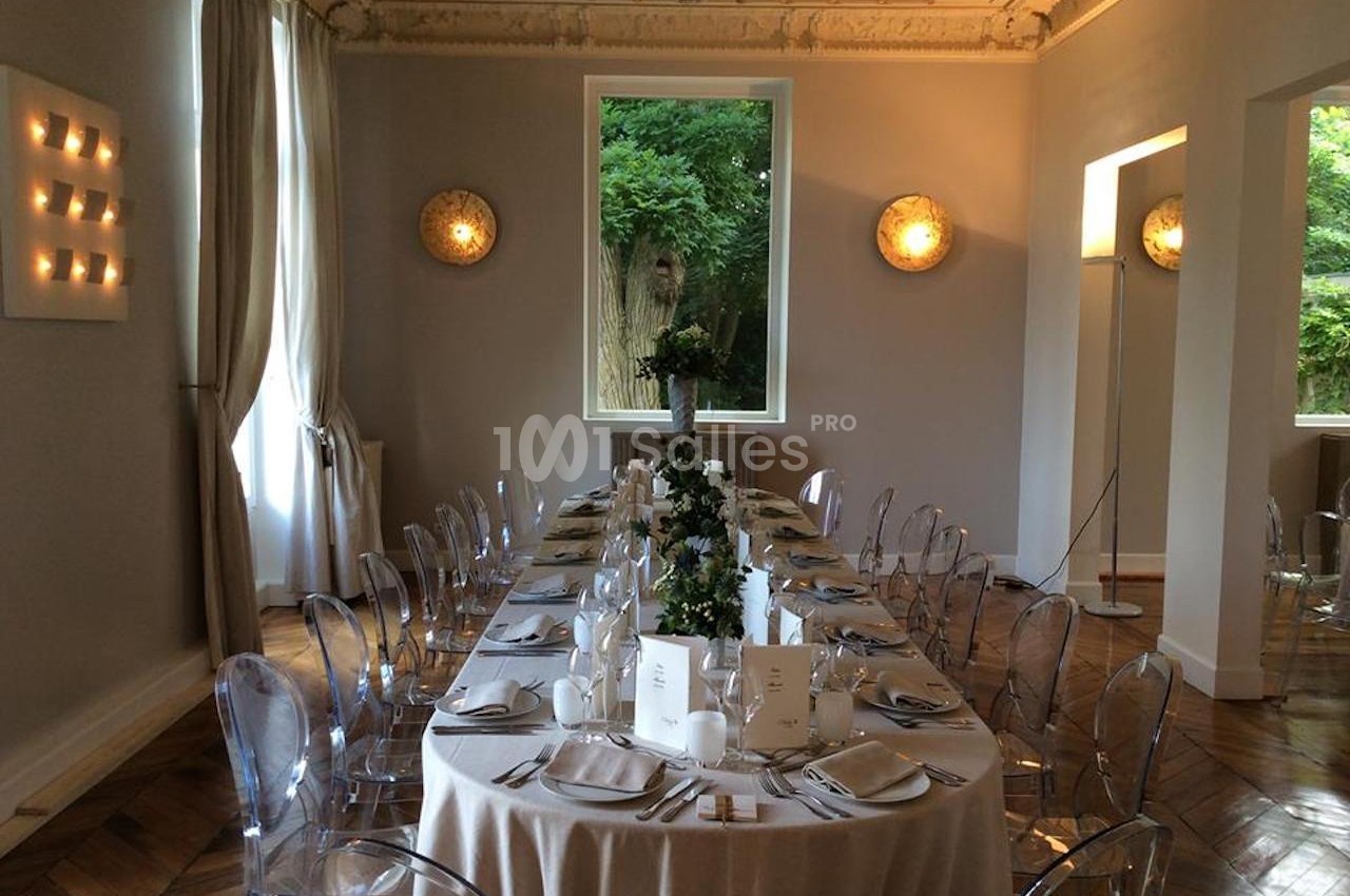 Salle élégante avec une grande table dressée, nappes blanches, chaises transparentes et vue sur un jardin verdoyant.