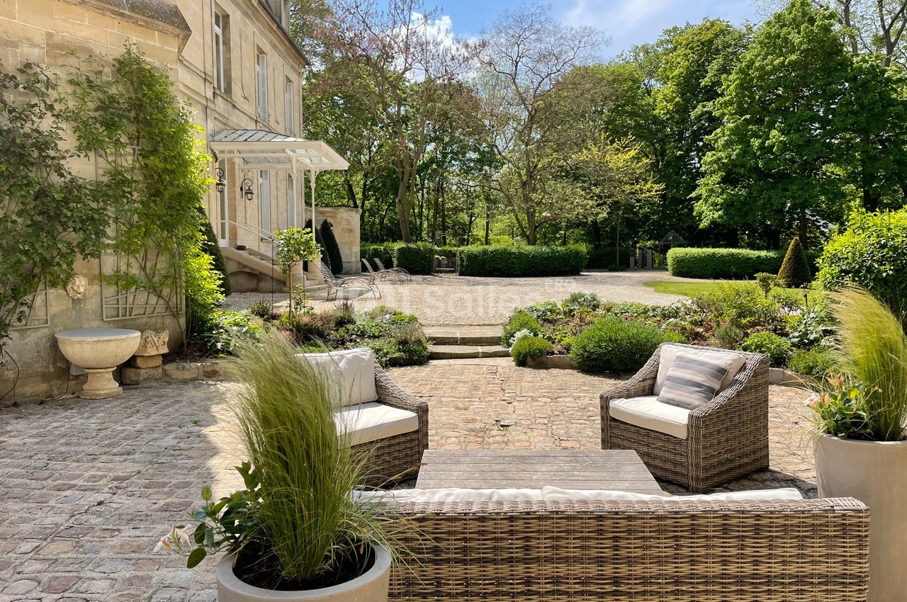Terrasse pavée avec mobilier en rotin, plantes en pot et vue sur un jardin arboré devant une maison en pierre.