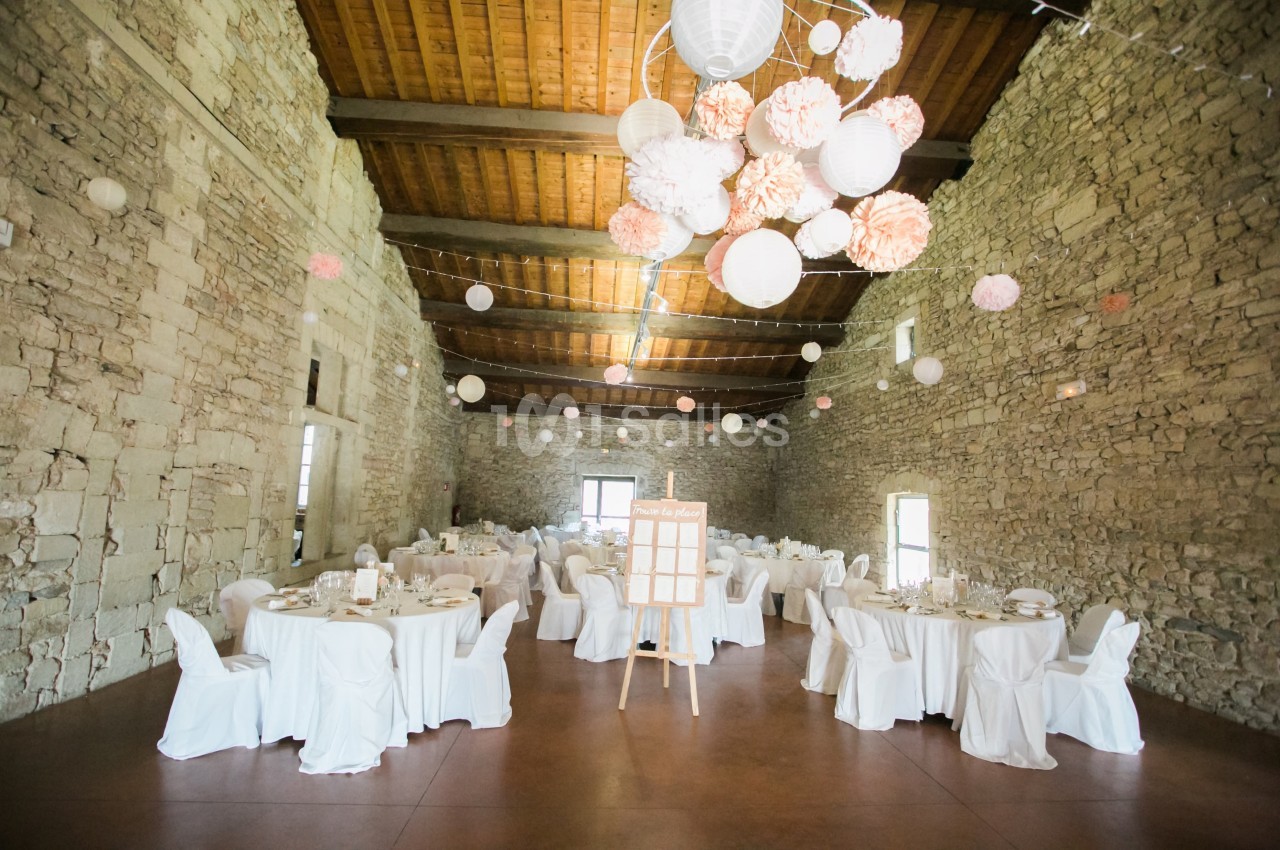 Salle en pierre avec tables rondes dressées pour un événement, décorée de lanternes et pompons suspendus au plafond.