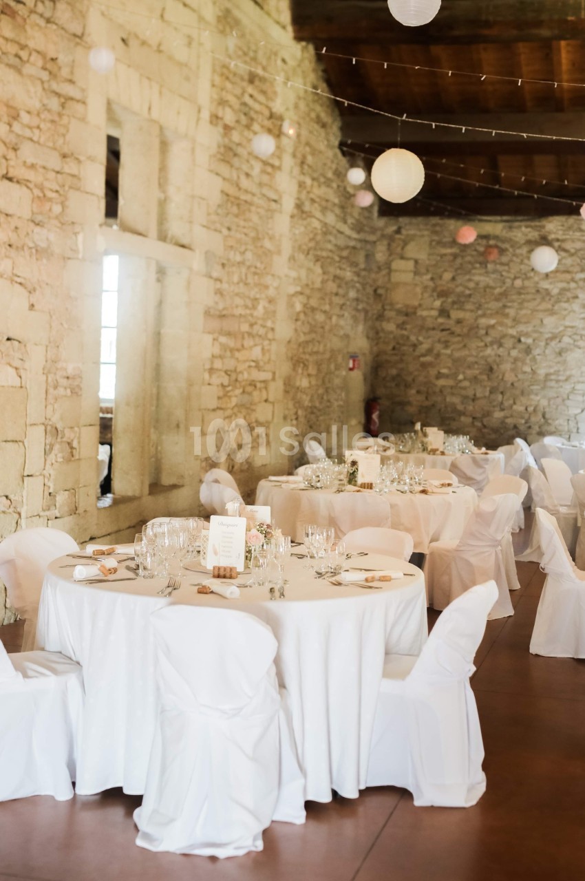Salle en pierre décorée pour un événement, avec des tables rondes dressées et des chaises couvertes de housses blanches.