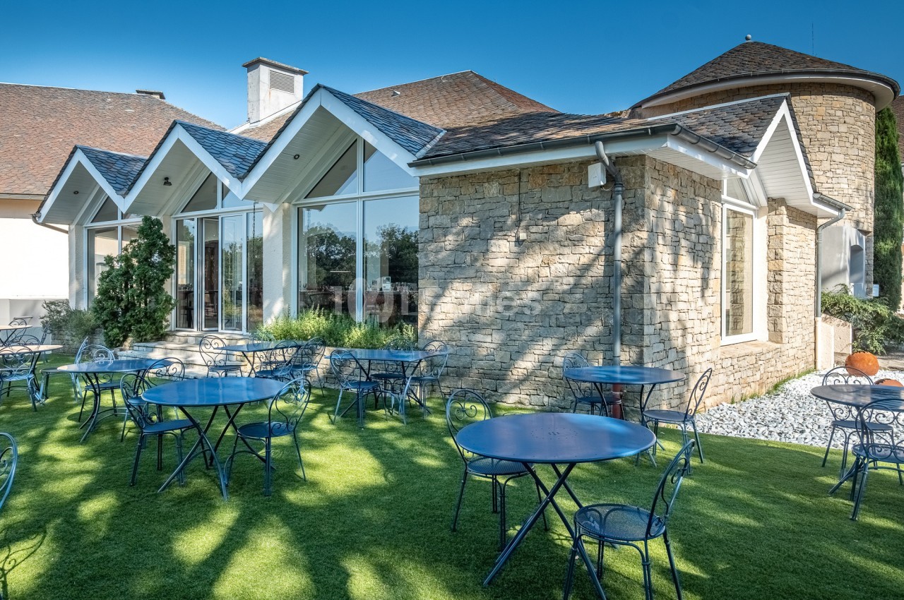 Terrasse avec tables et chaises en métal bleu sur pelouse, devant un bâtiment en pierre avec grandes baies vitrées.