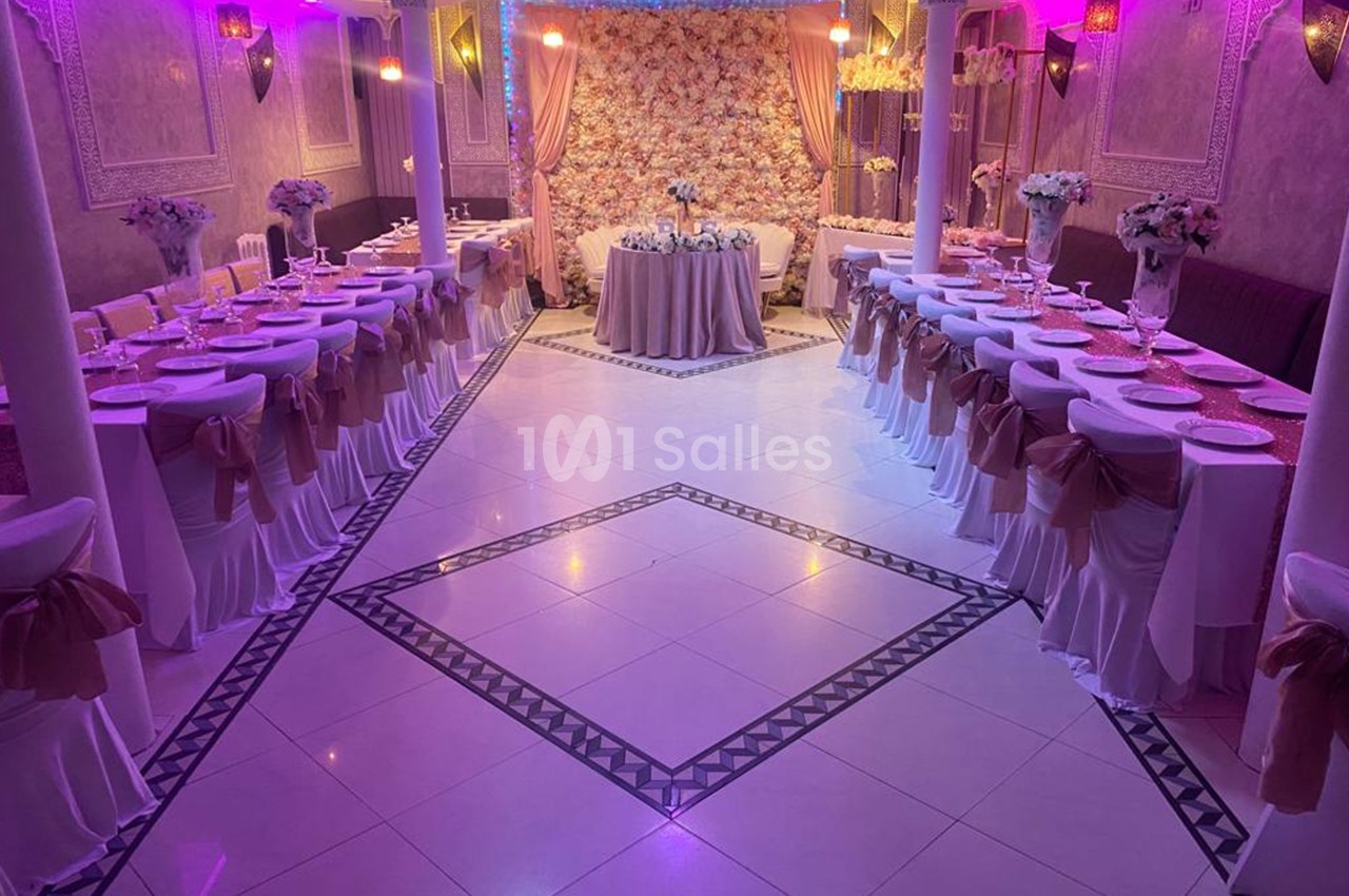 Salle de réception décorée avec des tables alignées, nappes blanches, chaises ornées de rubans et un mur floral en arrière…