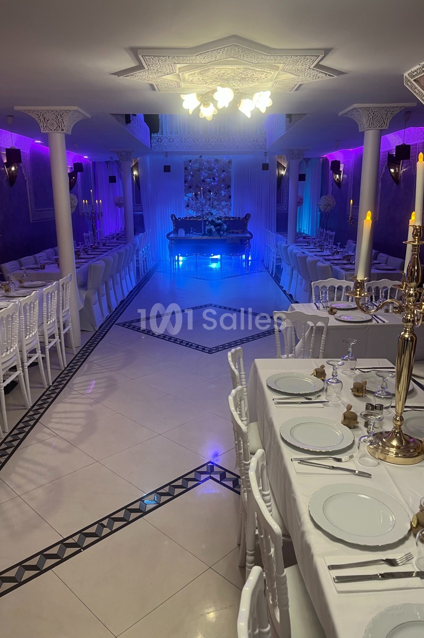 Salle de réception élégante avec tables dressées, chandeliers dorés et éclairage d'ambiance violet et bleu.