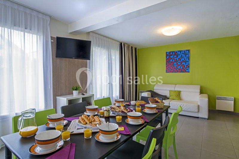 Table dressée pour un petit-déjeuner avec viennoiseries, jus d'orange et vaisselle colorée dans une salle lumineuse.