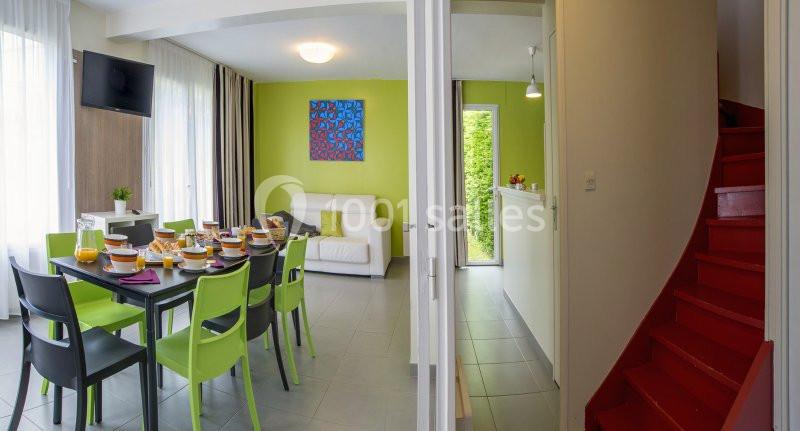 Salle à manger lumineuse avec table dressée, chaises vertes, canapé blanc, escalier rouge et vue sur un jardin.