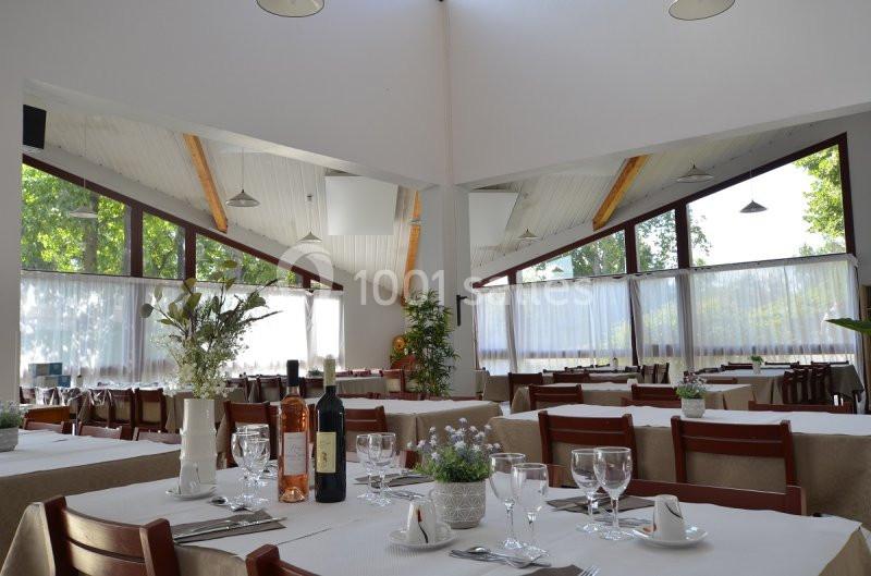 Salle de restaurant lumineuse avec tables dressées, nappes blanches, verres, bouteilles de vin et grandes baies vitrées.