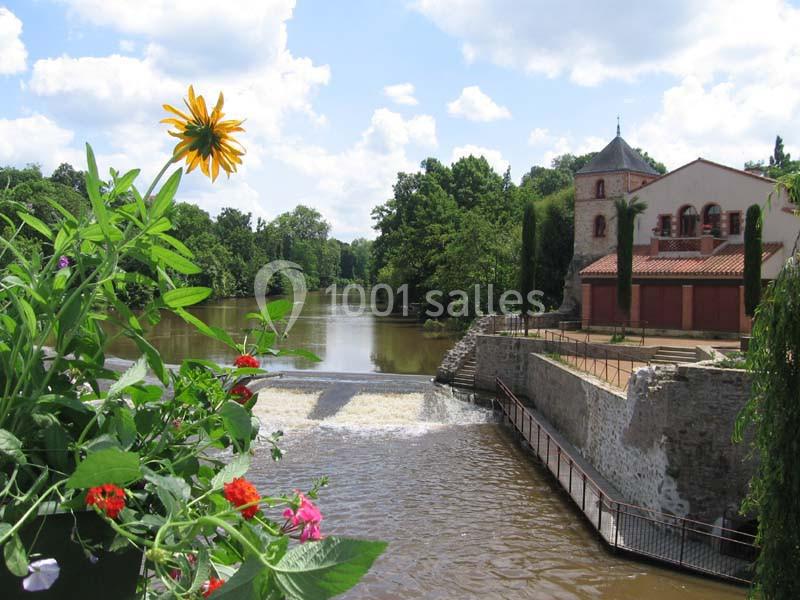 Vue d'une rivière avec un petit barrage, des fleurs au premier plan et un bâtiment en pierre entouré de verdure.