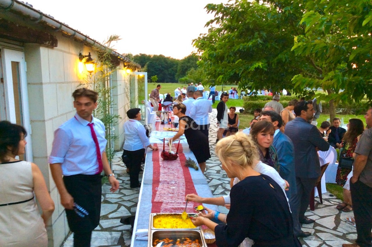 Des convives se servent à un buffet en extérieur, près d'un bâtiment, lors d'un événement en fin de journée.