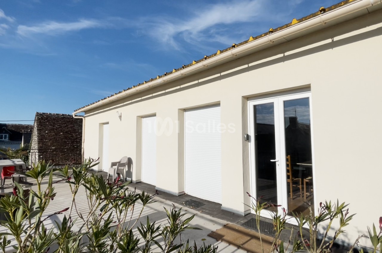 Façade d'une maison avec murs clairs, volets roulants fermés et terrasse entourée de végétation sous un ciel bleu.