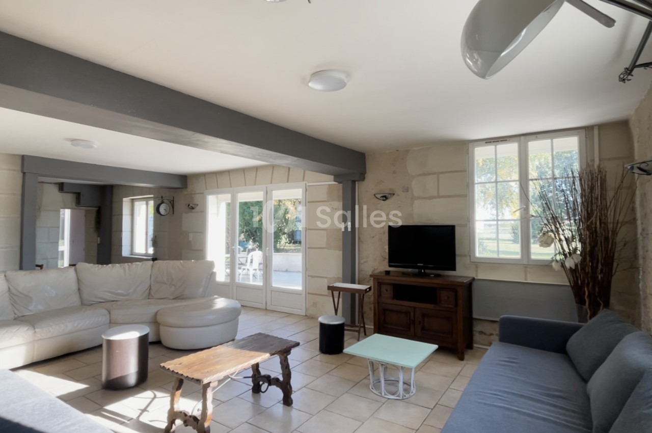 Salon lumineux avec canapé d'angle, table basse en bois, télévision sur meuble et grandes fenêtres donnant sur un jardin.