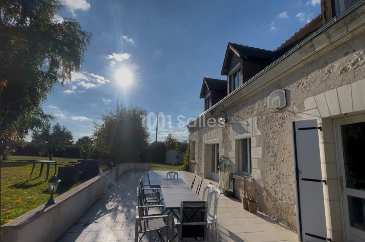 Terrasse ensoleillée avec une grande table et des chaises, adossée à une maison en pierre sous un ciel dégagé.