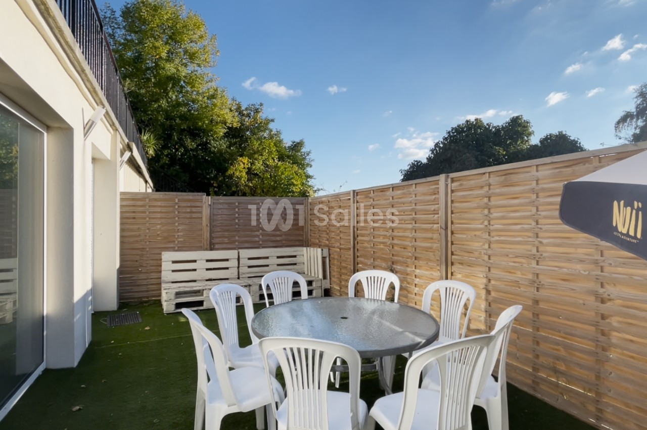 Terrasse extérieure avec table ronde en verre entourée de chaises blanches en plastique, clôturée par des panneaux en bois.