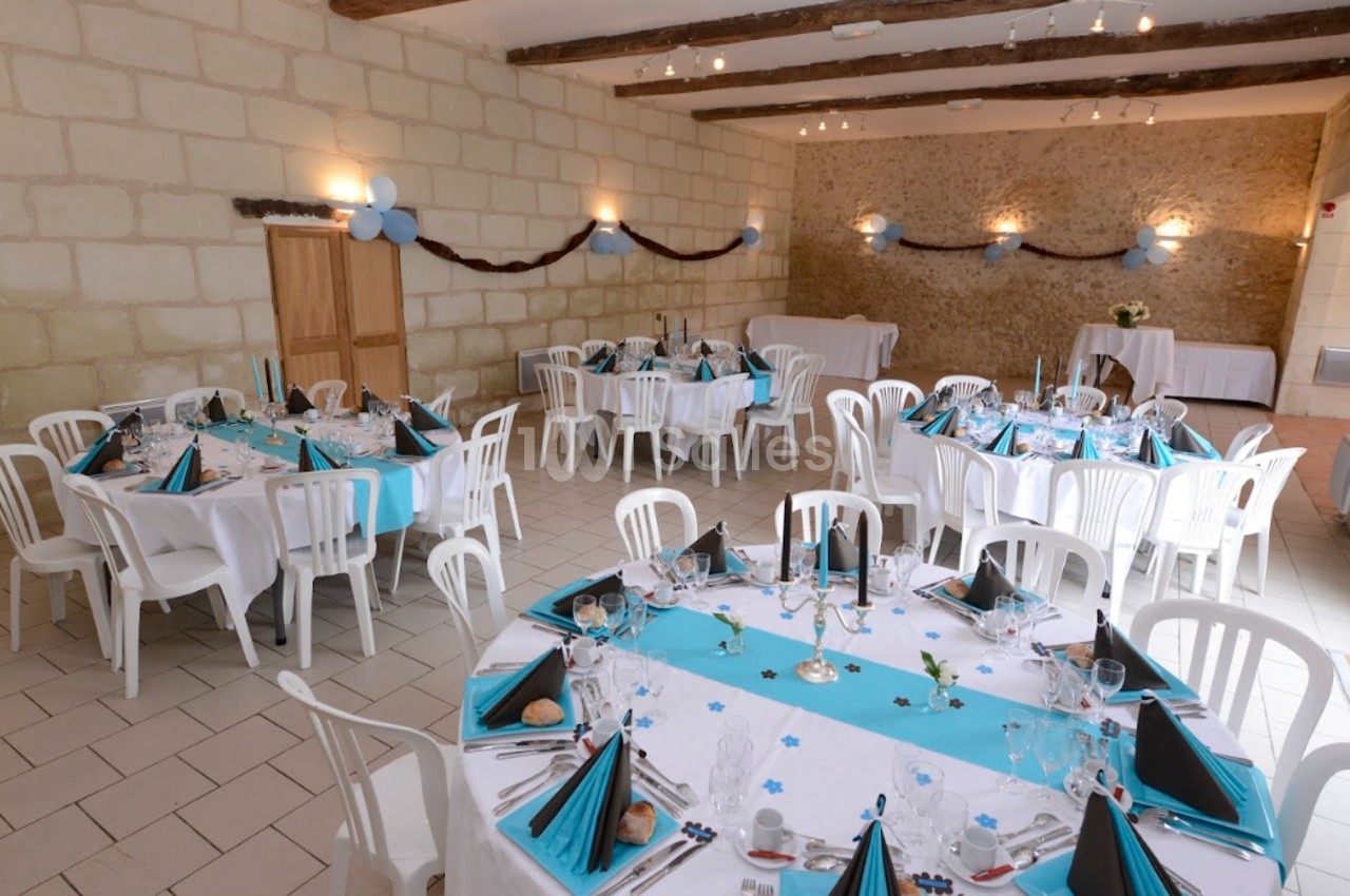 Salle décorée pour un événement avec tables rondes dressées, nappes blanches et bleues, chaises blanches et guirlandes…