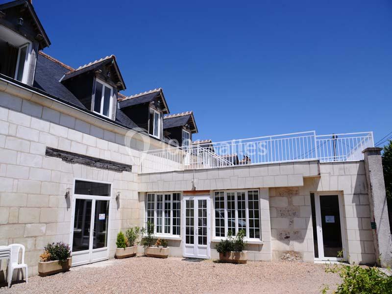 Façade d'une maison en pierre avec fenêtres blanches, terrasse à l'étage et cour gravillonnée.
