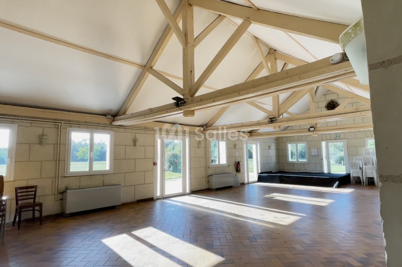 Salle lumineuse avec charpente apparente, parquet en bois, grandes fenêtres et empilement de chaises dans un coin.