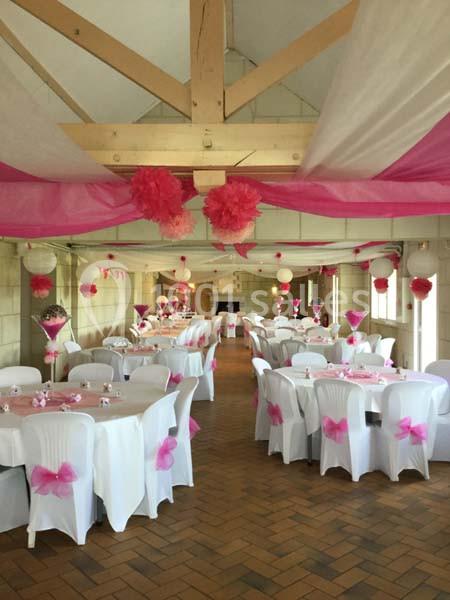 Salle décorée pour un événement avec nappes blanches, chaises ornées de nœuds roses et guirlandes suspendues.