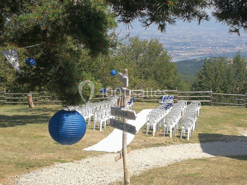 Chaises blanches alignées en extérieur pour une cérémonie, avec lanternes bleues et vue sur un paysage vallonné.