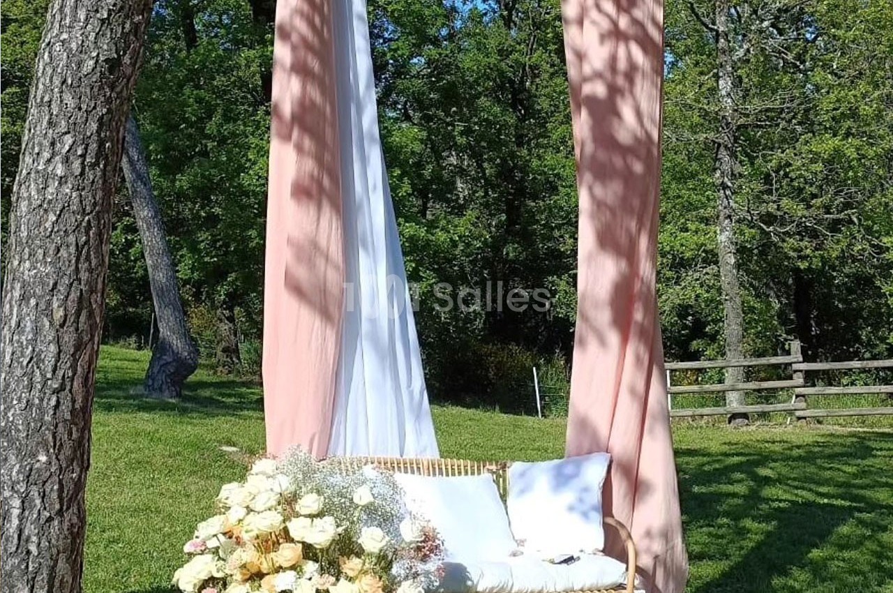 Chaise en rotin décorée de fleurs blanches et roses, placée sous des voilages suspendus entre deux arbres dans un jardin.