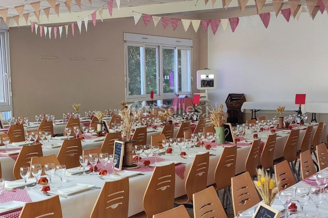 Salle décorée pour un repas festif avec tables dressées, nappes blanches, chemins de table rouges et guirlandes suspendues.