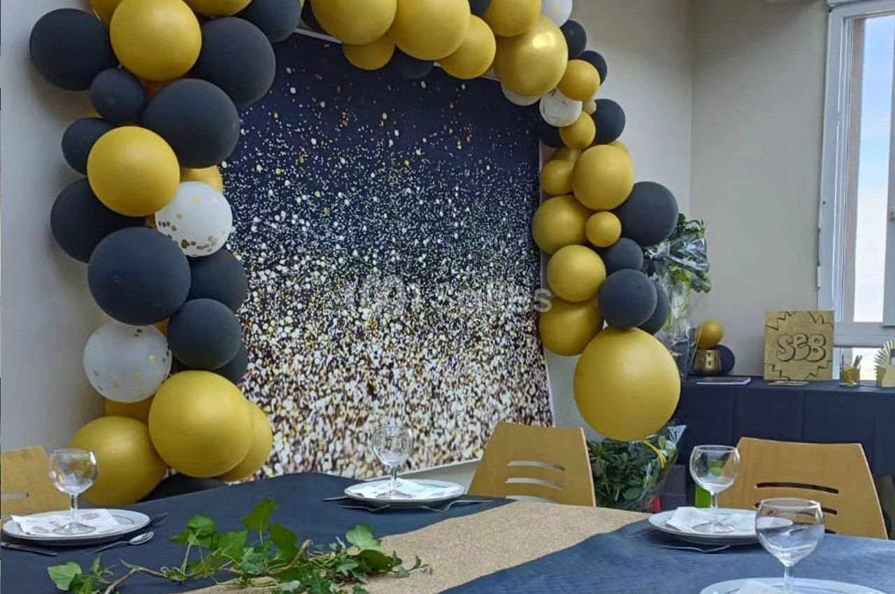 Décoration de salle avec arche de ballons noirs et dorés, table dressée et fond scintillant pour un événement.