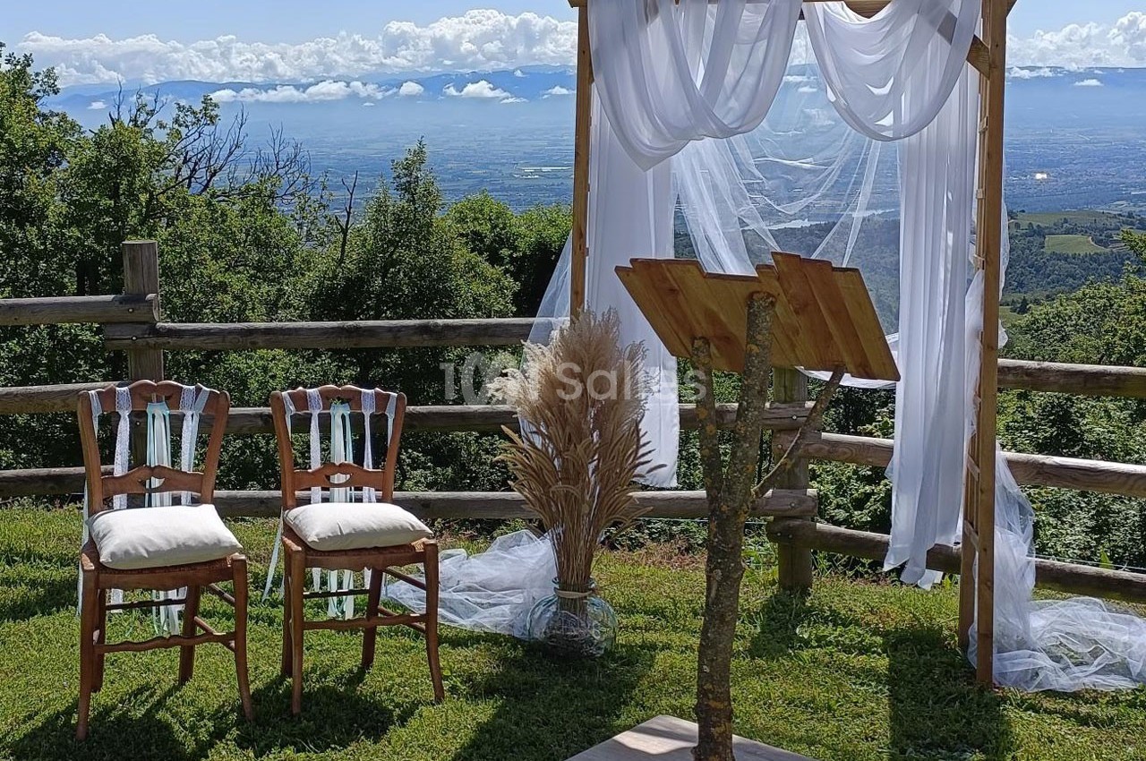 Arche en bois décorée de voilages blancs, deux chaises et pupitre sur une pelouse avec vue sur un paysage vallonné.