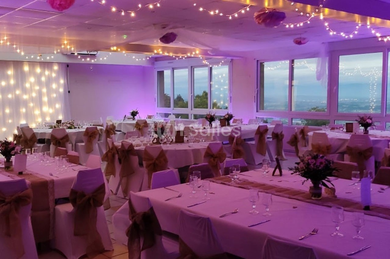 Salle de réception décorée avec des guirlandes lumineuses, tables dressées et vue sur un paysage extérieur.