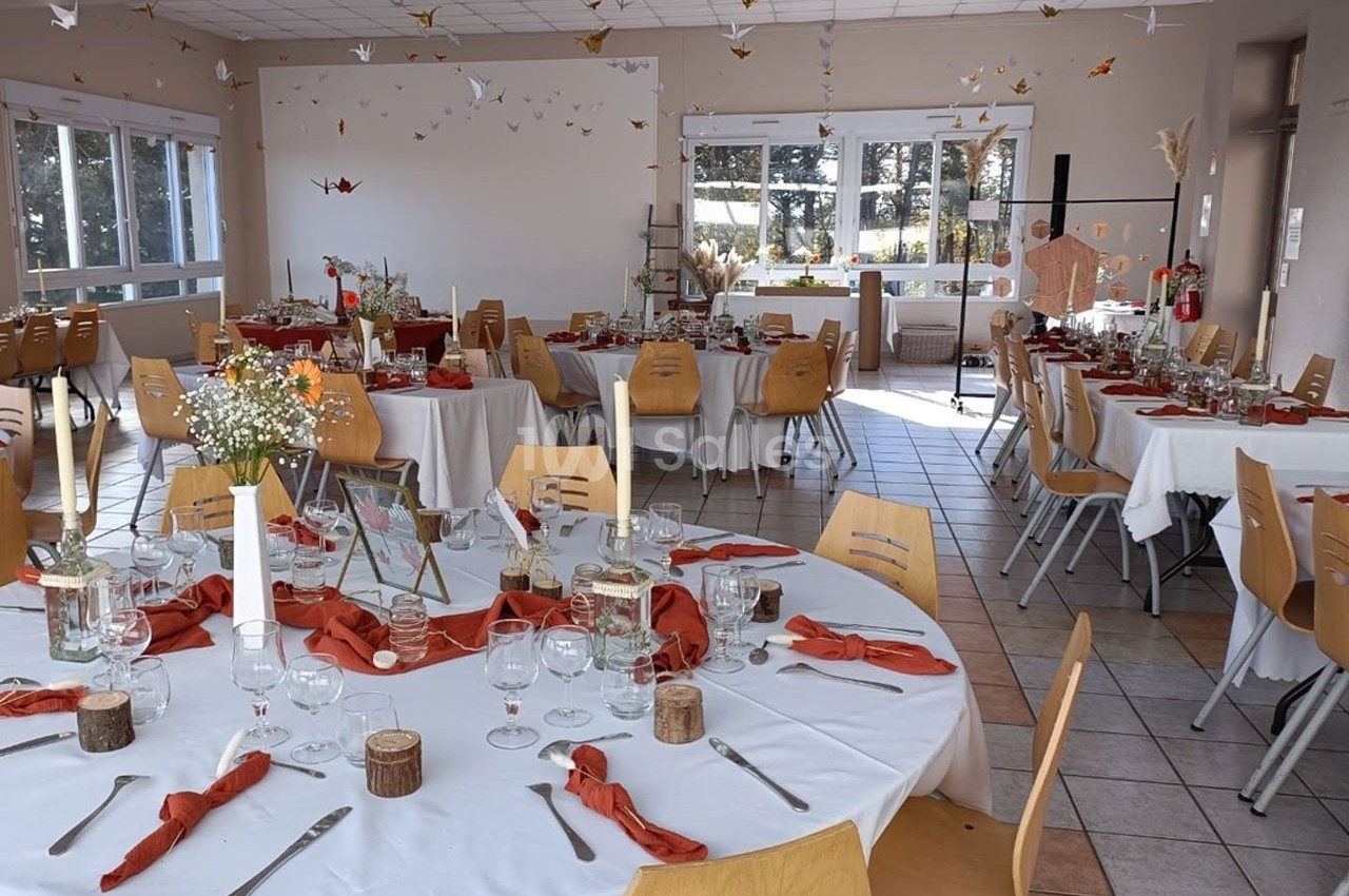 Salle décorée pour un événement avec tables rondes dressées, nappes blanches, serviettes orange et pliages en papier…