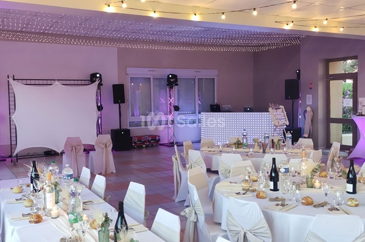 Salle de réception décorée pour un événement, avec tables dressées, chaises ornées et éclairage festif.