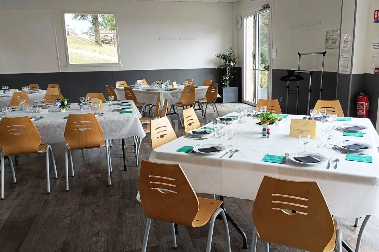 Salle de réception lumineuse avec des tables dressées, nappes blanches, chaises en bois et vue sur l'extérieur.