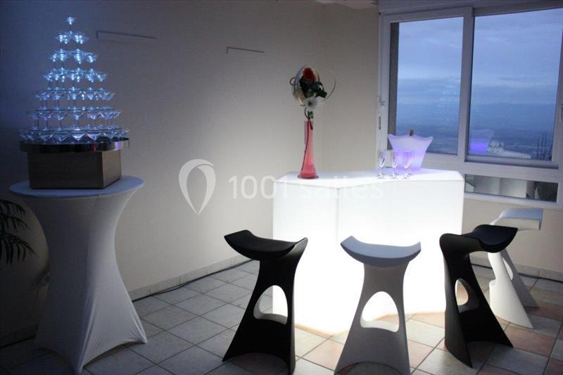 Bar lumineux avec tabourets noirs et blancs, table haute avec une pyramide de verres et vase contenant une rose.