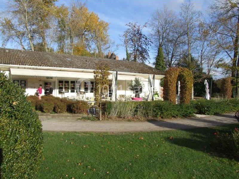 Bâtiment blanc entouré de verdure avec terrasse, parasols fermés et arbres en arrière-plan par temps ensoleillé.