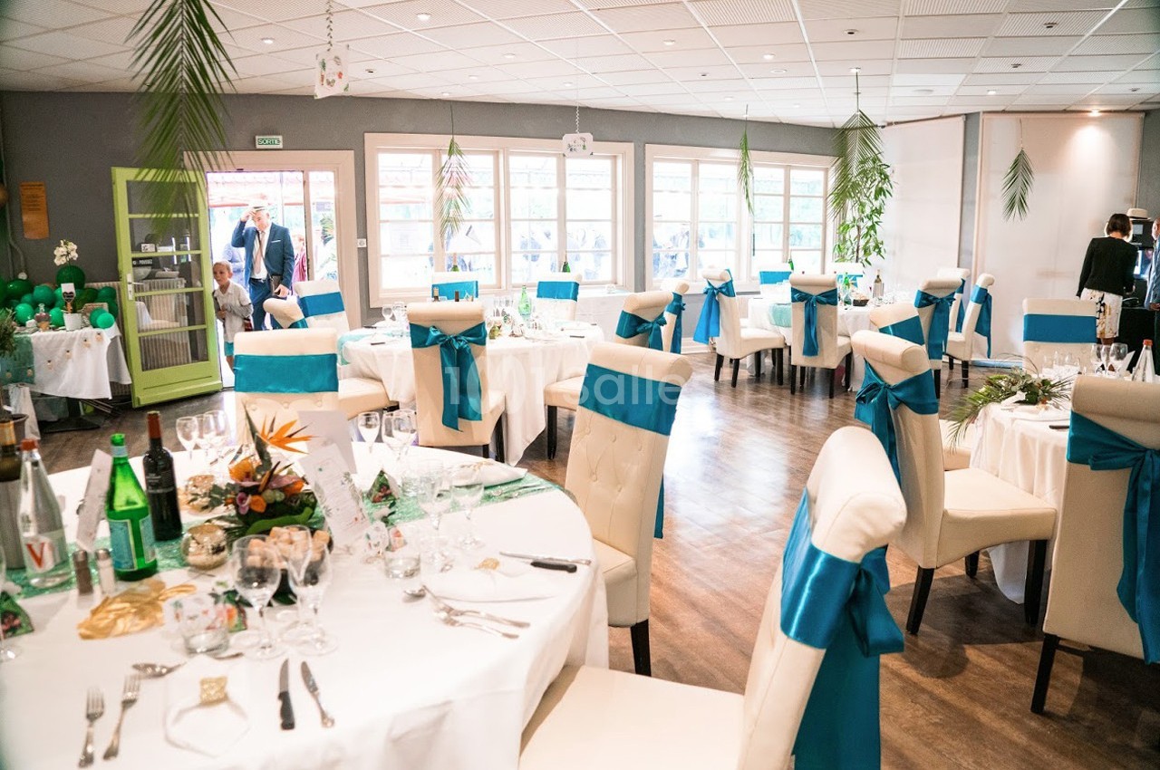 Salle de réception décorée avec des tables rondes, nappes blanches, chaises ornées de nœuds bleus et lumière naturelle.