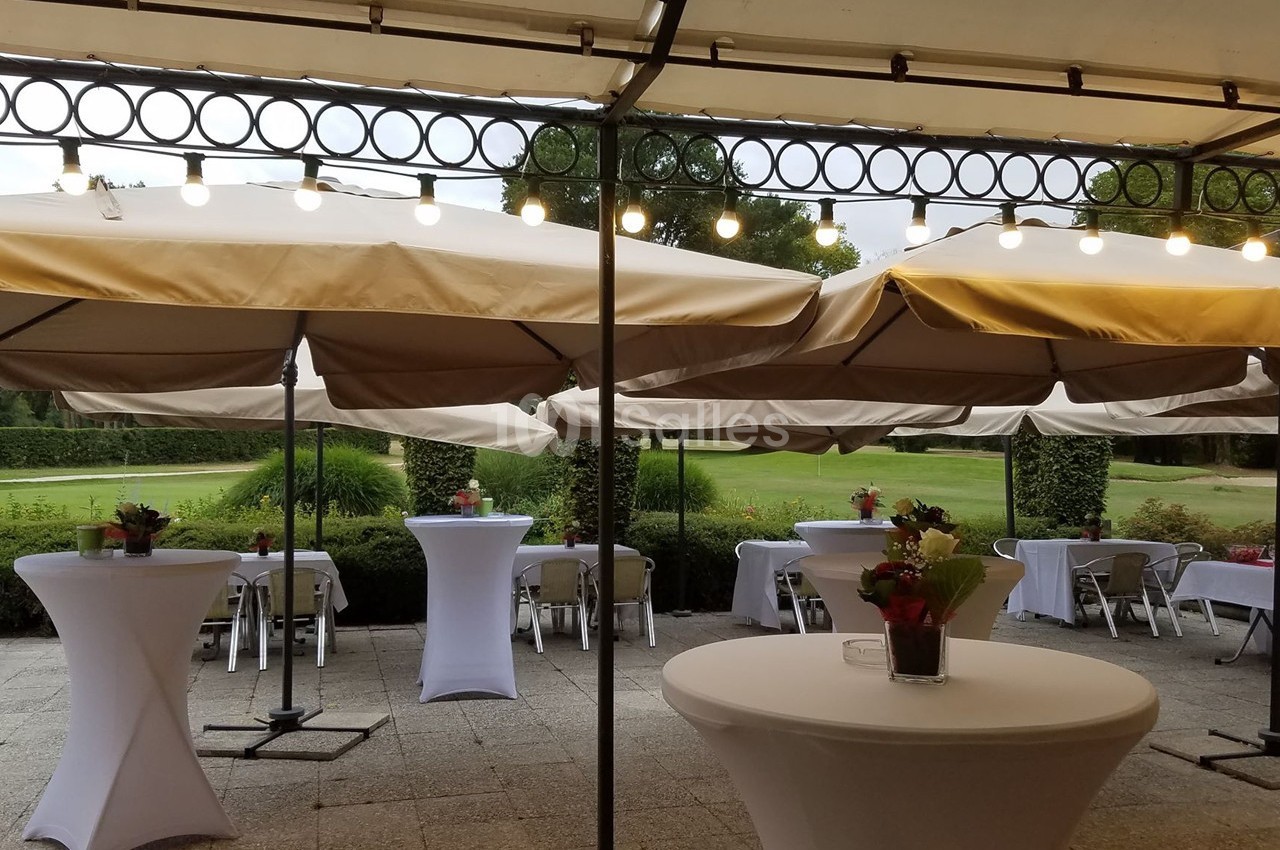 Terrasse aménagée avec tables hautes, parasols beiges et guirlandes lumineuses, donnant sur un jardin verdoyant.