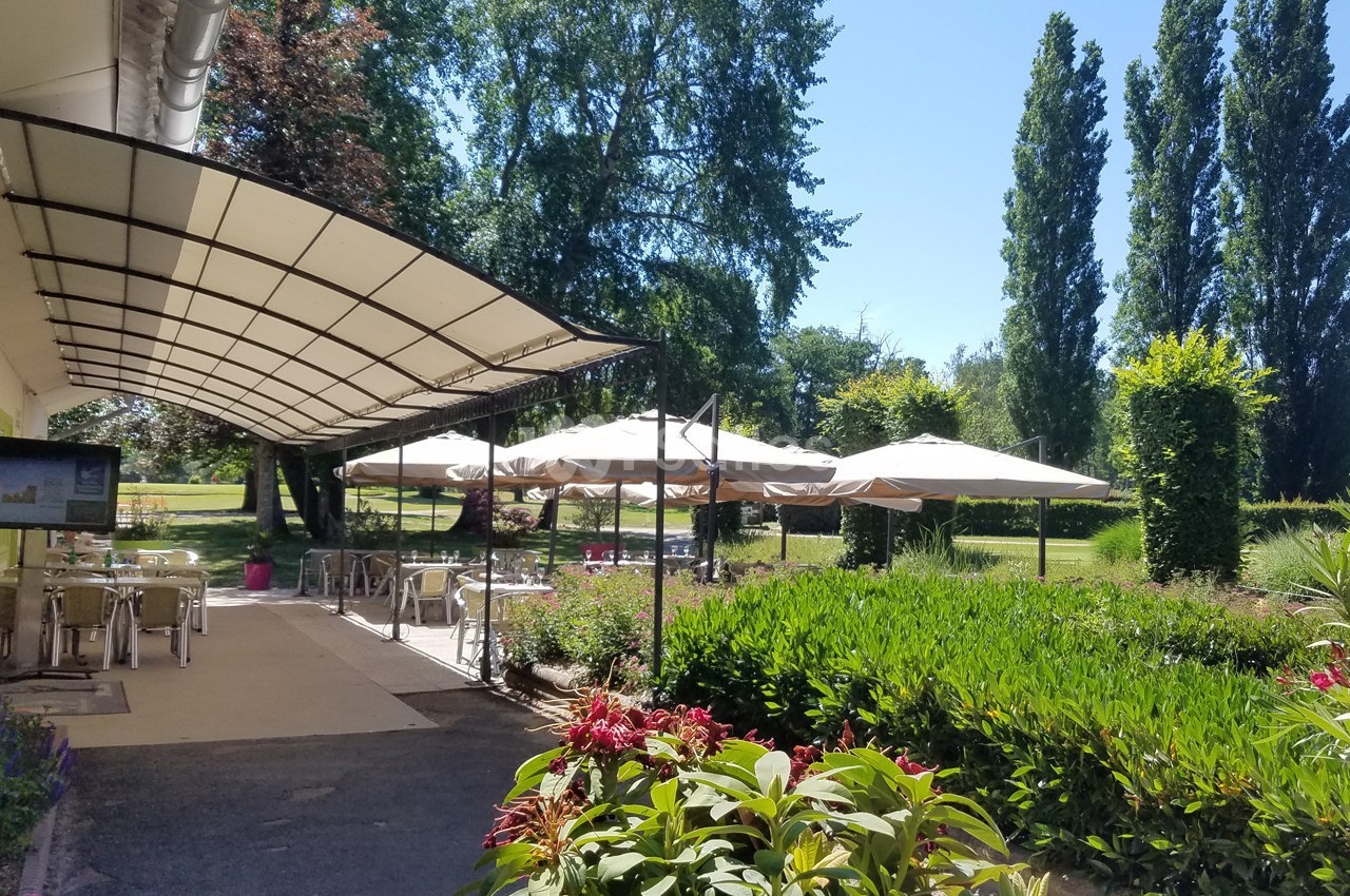 Terrasse ombragée avec tables et chaises, entourée de verdure et de fleurs, sous un ciel dégagé.
