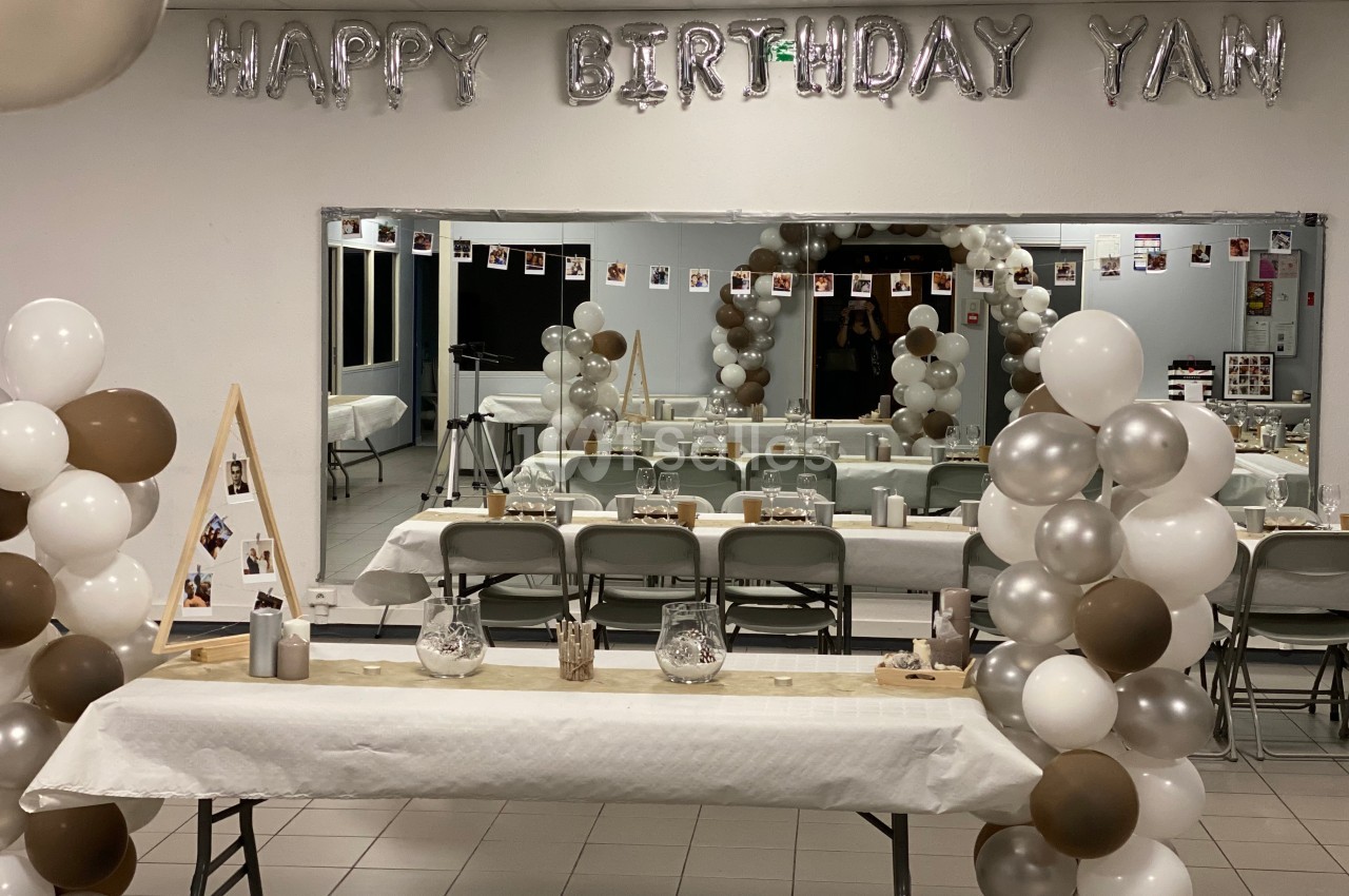 Salle décorée pour un anniversaire avec ballons blancs, marron et argentés, table dressée et guirlande ’Happy Birthday Yan’.