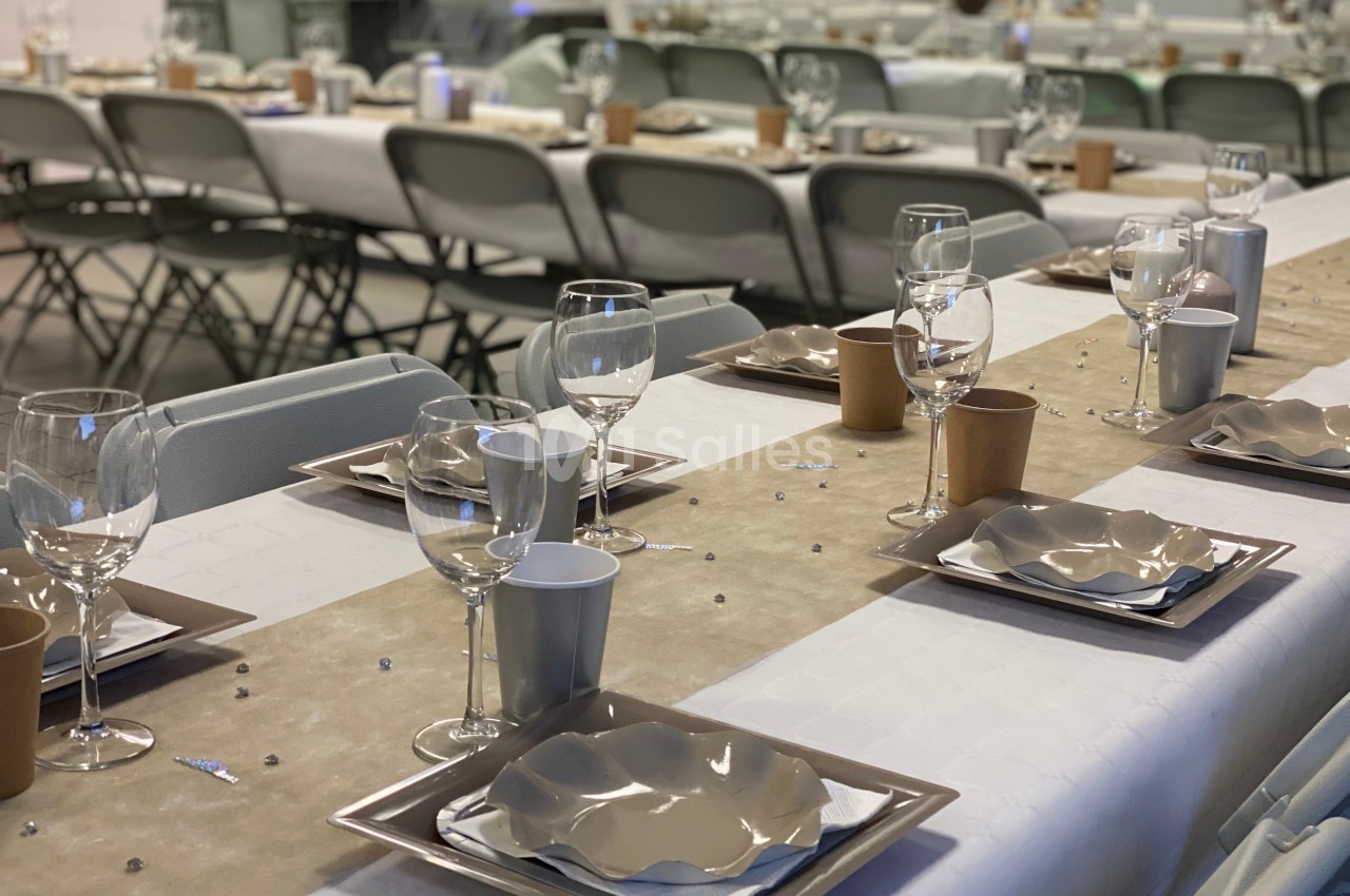 Tables dressées avec nappes beige et blanche, assiettes modernes, verres à pied et chaises pliantes dans une salle.