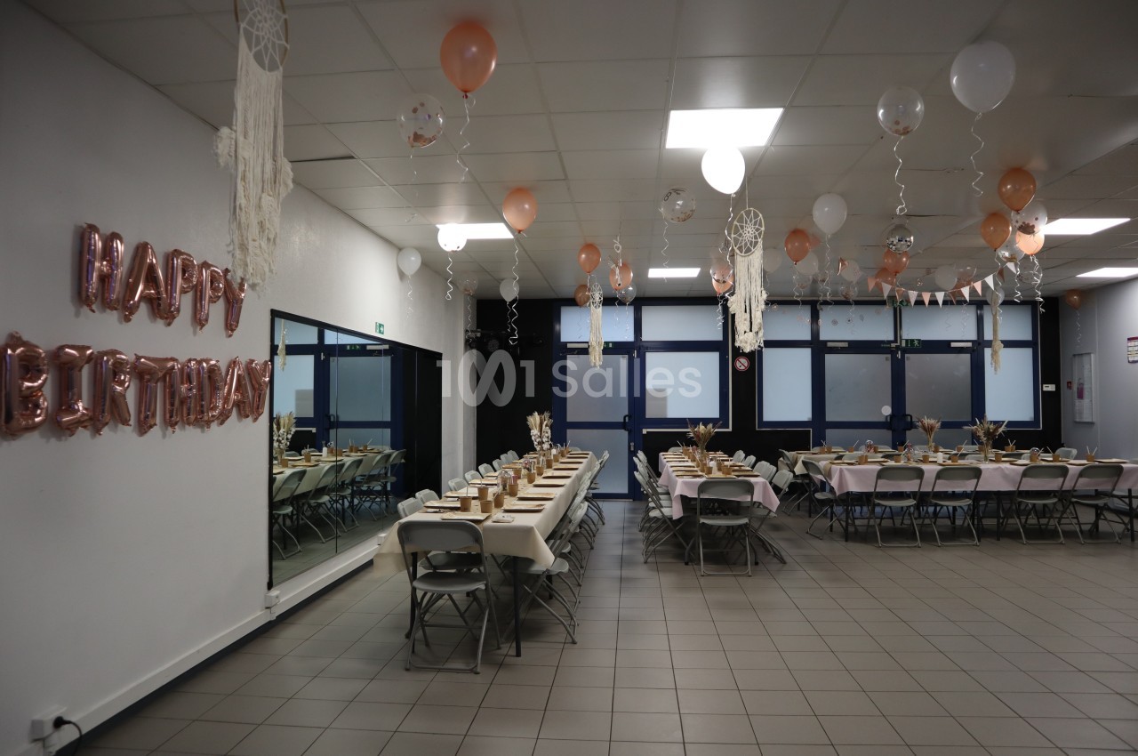 Salle décorée pour un anniversaire avec tables dressées, ballons suspendus et guirlandes dans des tons pastel.