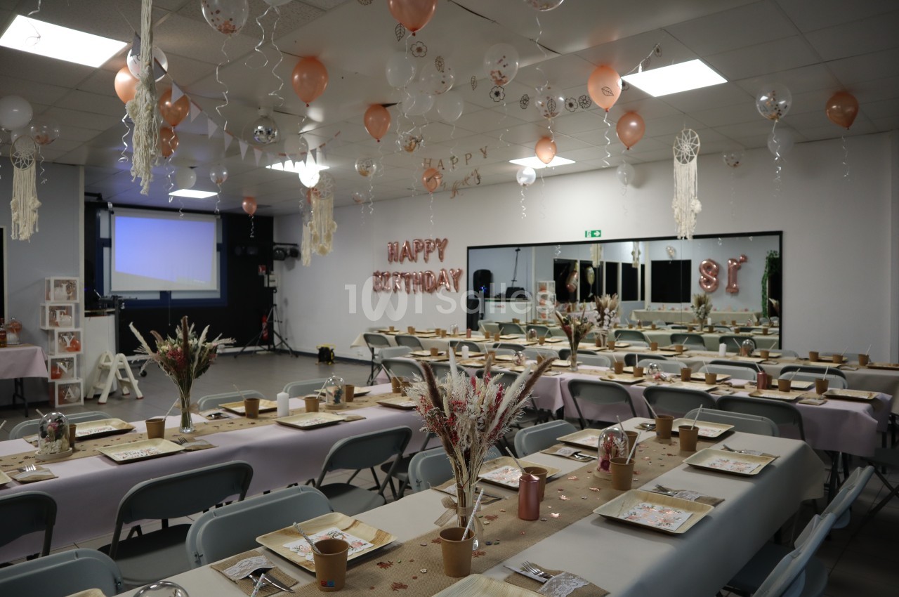 Salle décorée pour un anniversaire avec tables dressées, ballons suspendus et projecteur en arrière-plan.