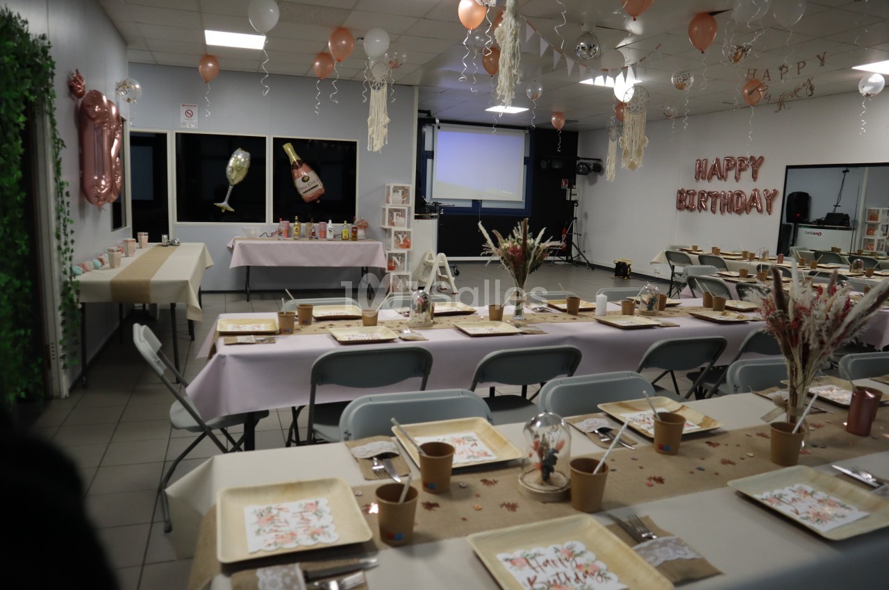 Salle décorée pour un anniversaire avec tables dressées, ballons suspendus et guirlande ’Happy Birthday’ au mur.