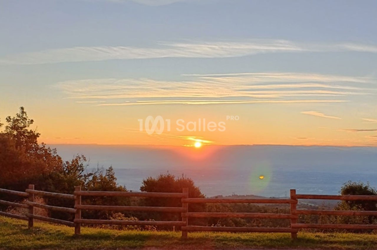 Lever de soleil sur un paysage vallonné, avec une clôture en bois au premier plan et un ciel partiellement nuageux.
