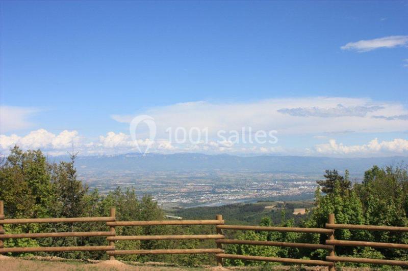 Vue panoramique sur une vallée avec des montagnes à l'horizon, bordée par une clôture en bois et des arbres.