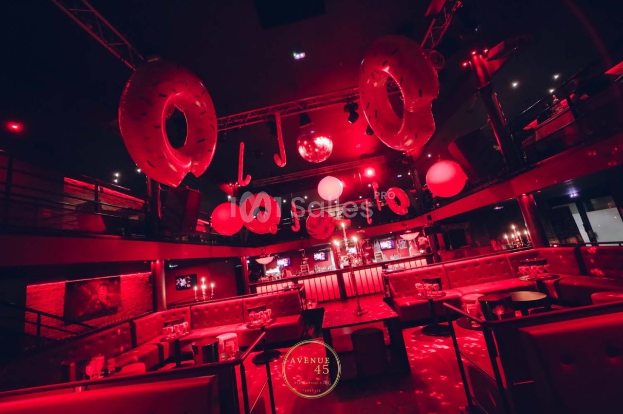 Salle de fête avec éclairage rouge, décorations suspendues et tables entourées de banquettes.