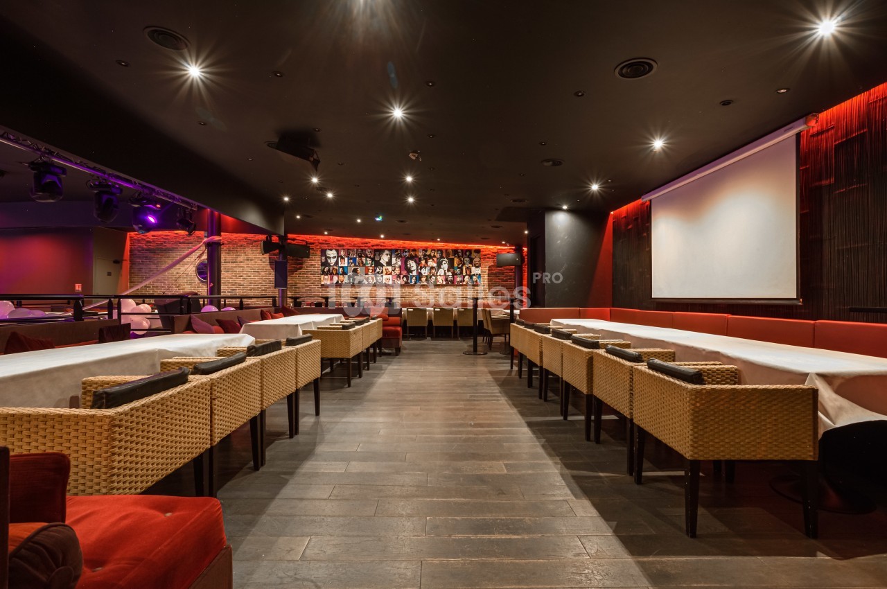 Salle de restaurant avec tables alignées, chaises en osier, éclairage tamisé et bar en arrière-plan.