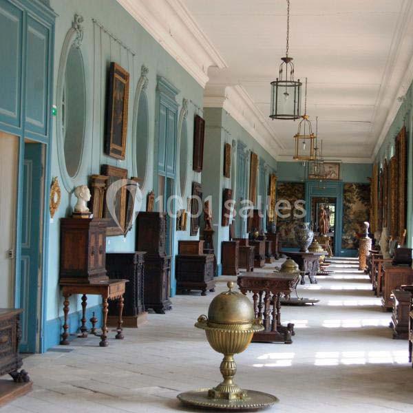 Galerie intérieure avec murs bleus, meubles anciens, tableaux et objets décoratifs alignés le long d'un couloir lumineux.