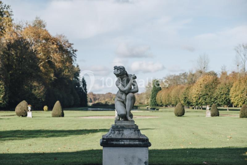 Statue en pierre représentant une figure assise dans un parc verdoyant avec des arbres aux couleurs automnales.