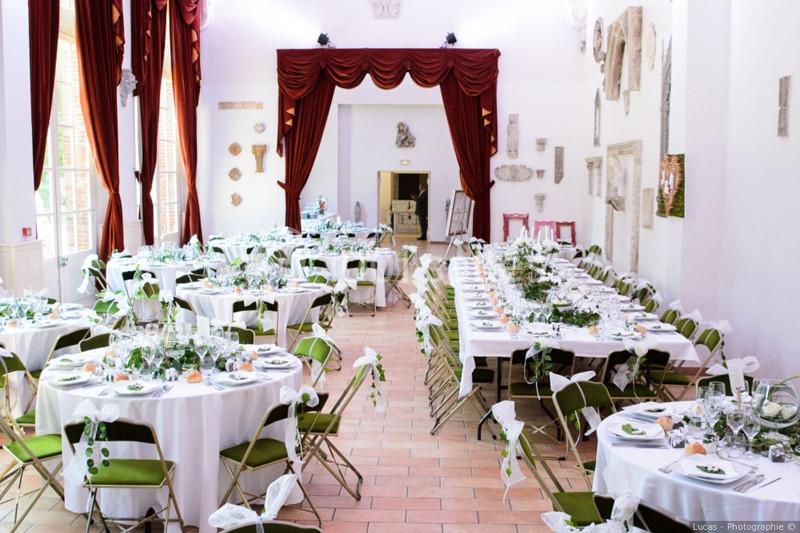 Salle de réception lumineuse avec tables décorées pour un événement, chaises pliantes et rideaux rouges en arrière-plan.
